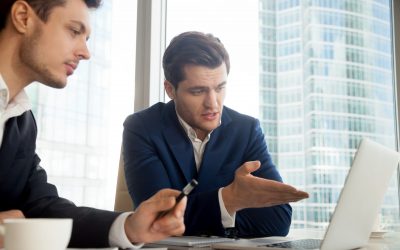 Handsome businessman sitting at desk with partner and pointing on laptop screen when explaining project advantages. Business leaders discussing details of companies collaboration. CEO presenting offer